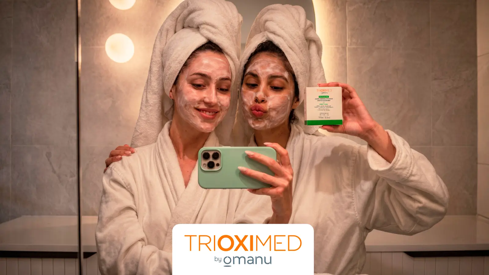 Dos mujeres con mascarilla facial y bata de baño realizan su rutina de cuidado de la piel frente a un espejo en un entorno de spa.