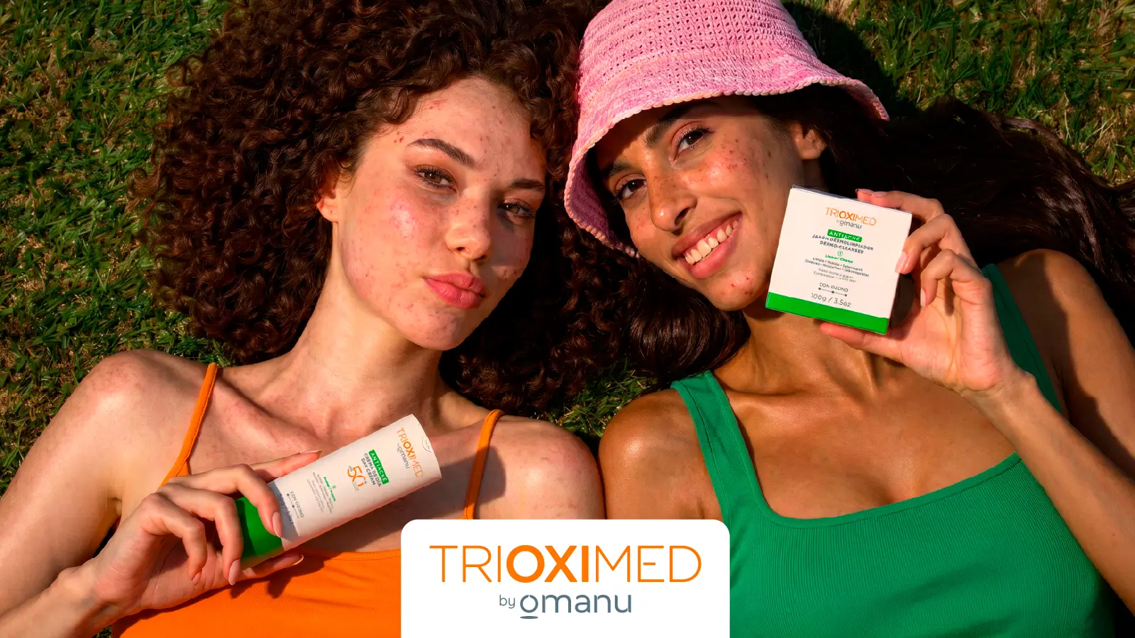 Dos mujeres con piel acneica recostadas sobre césped sosteniendo productos antiacné Trioximed by Omanu para rutina de skincare y cuidado facial.