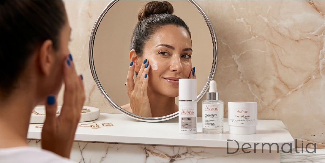Mujer aplicando crema facial frente al espejo junto a productos Avène DermAbsolu, Retrinal y Hyaluron Activ B3 para el cuidado antiedad.